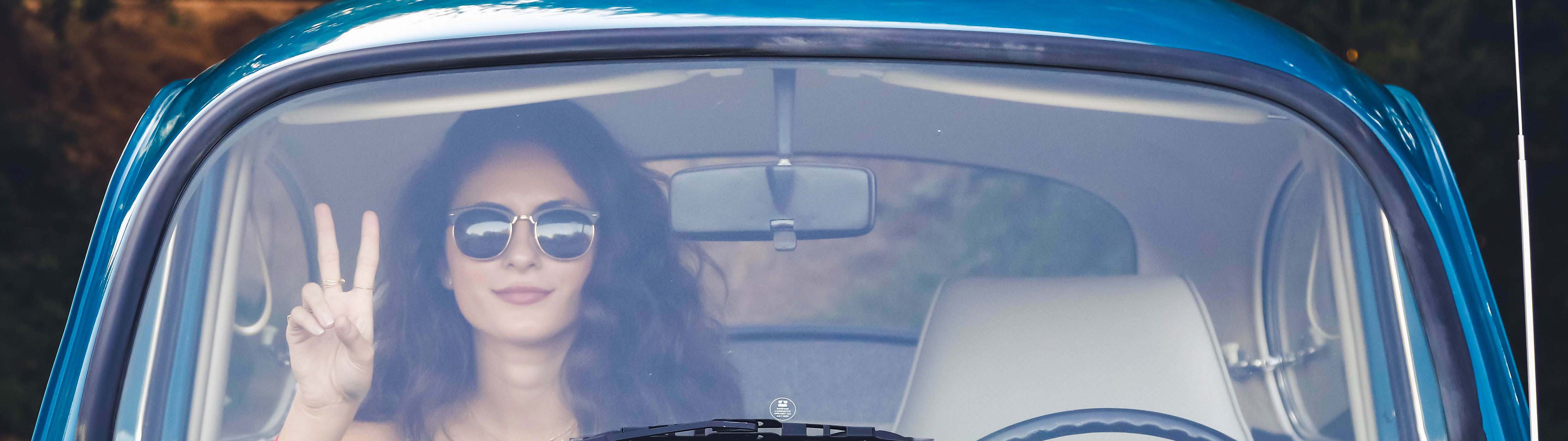 Photo of girl with brown hair and sunglasses sitting in a vintage blue Volkswagen bug. She’s giving the peace sign.